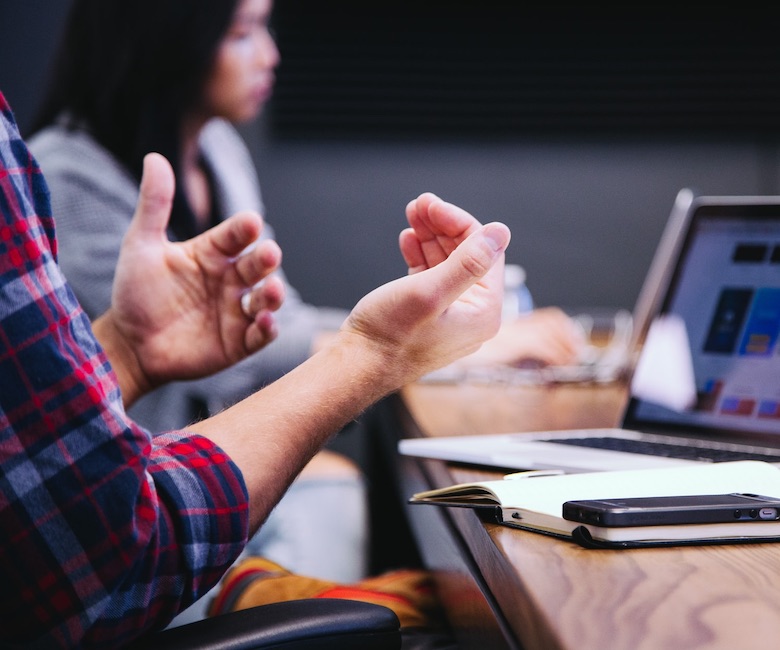 A team is in a meeting with their laptops open and notebooks out. One of the team members speaks emphatically, gesturing to his team.