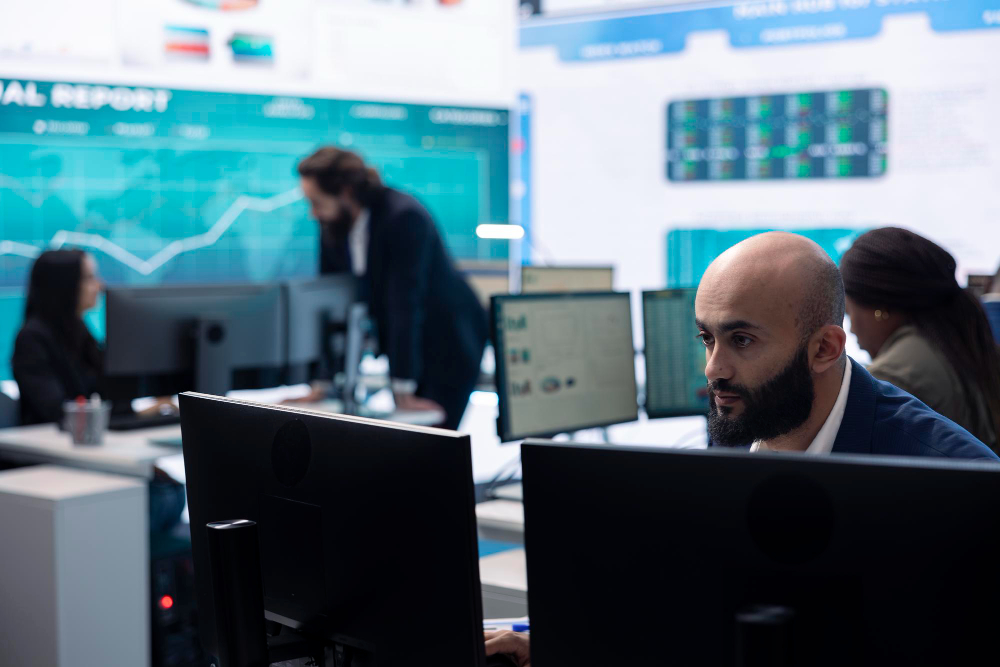 A man in a suit sits concentrated in front of two computer screens, while his colleagues work and converse in the background in front of a big graph. 