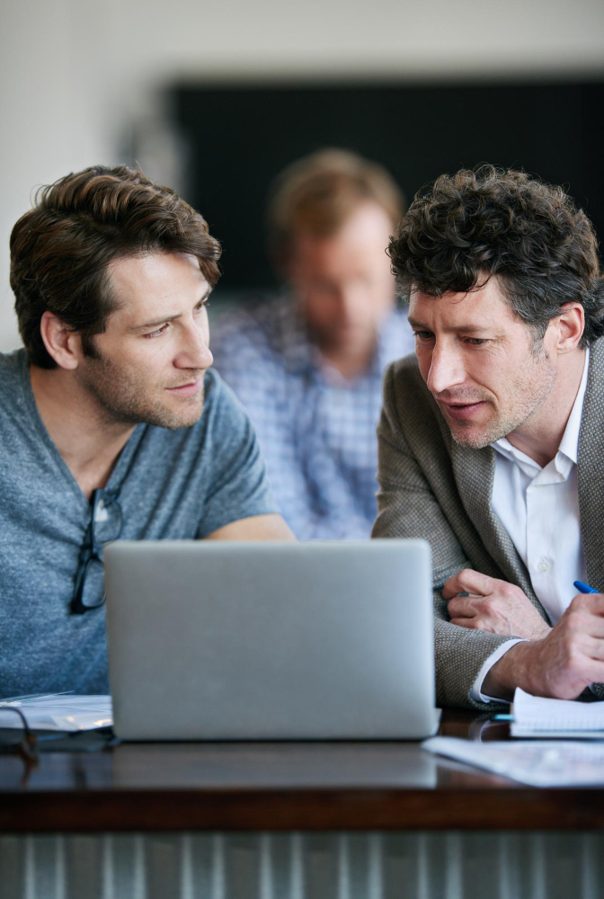 Two people are sitting at a desk together, looking at a laptop screen deep in thought. 