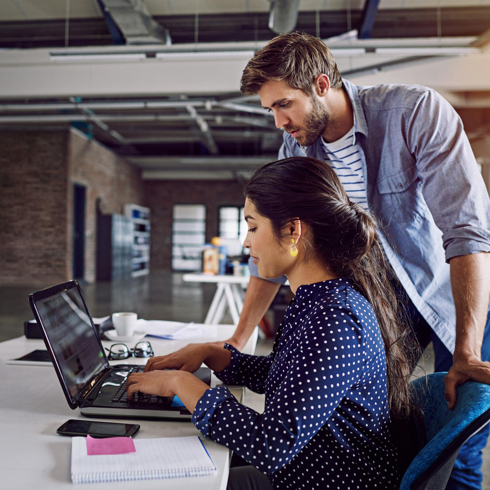 Two people are at a desk in a big open-concept office. One is sitting and typing on her laptop while another stands over her, examining her work. The desk has papers, notebooks, and coffee. 